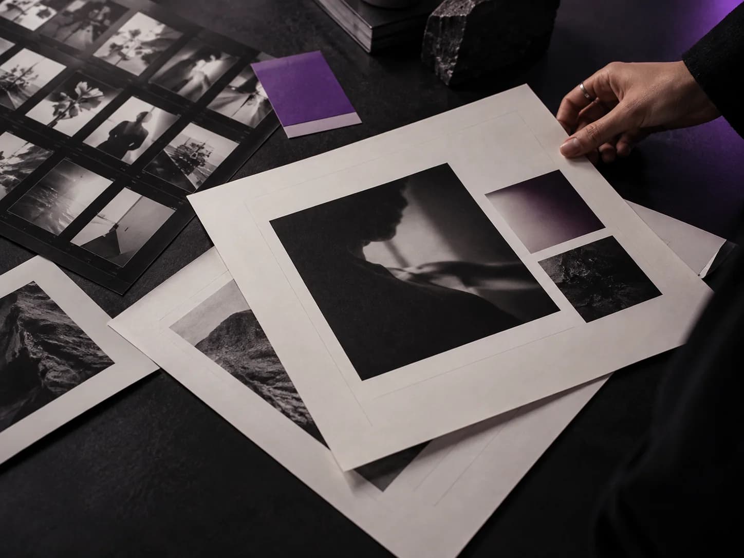 Hands arranging photographic and layout proofs on a dark creative worktable.