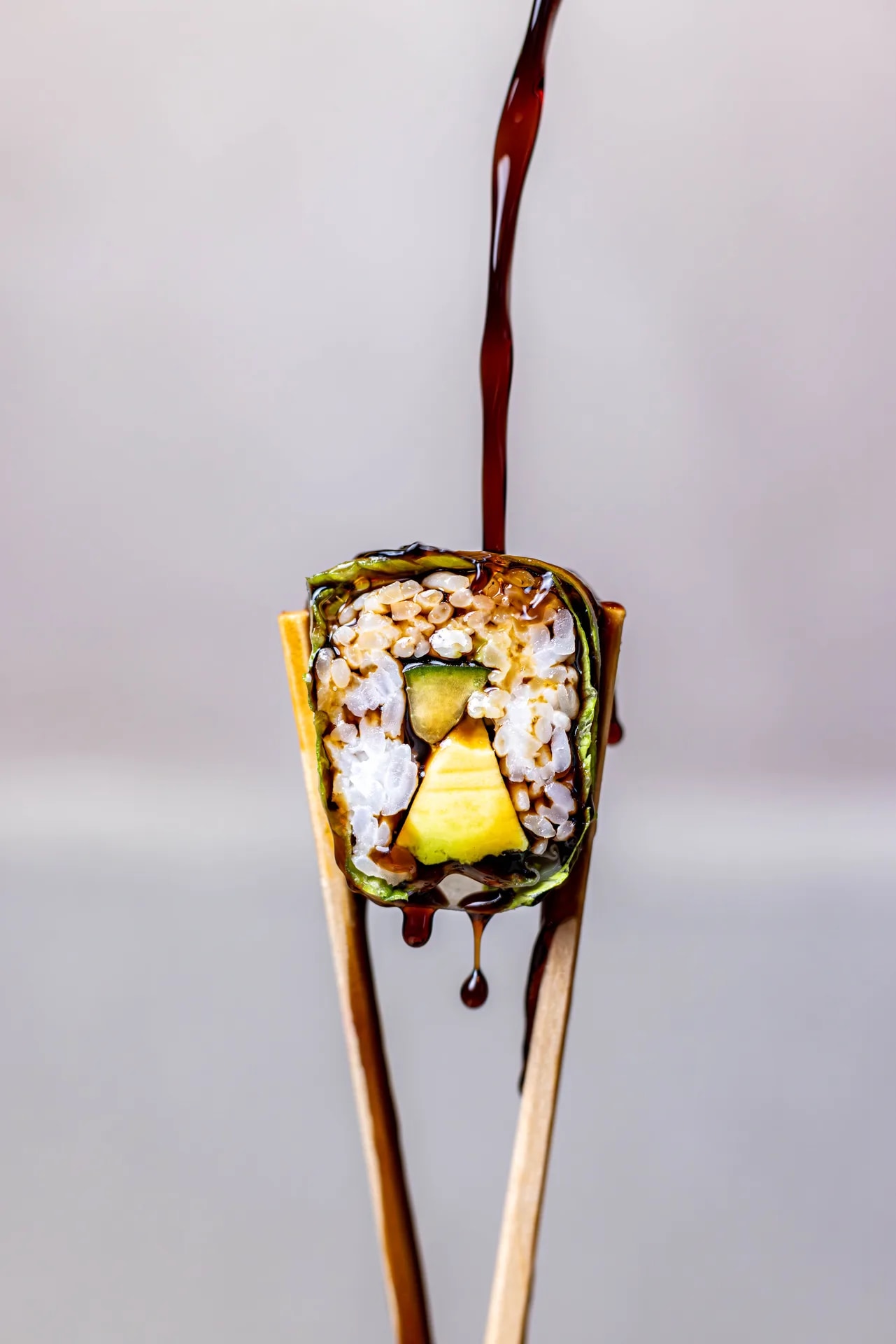 Sushi roll held between chopsticks while soy sauce is poured over it.